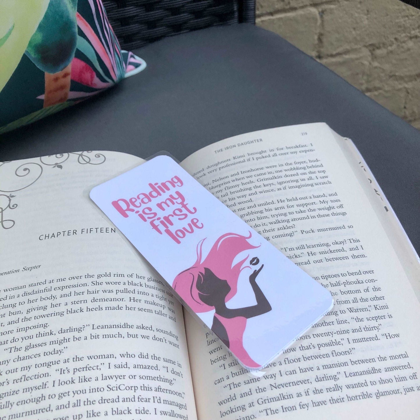 First Love Bookmark - Diamond Candlelight - girl with pink hair blowing a kiss, pink writing which says reading is my first kiss. Bookmark laying over a book sitting on a chair outside.