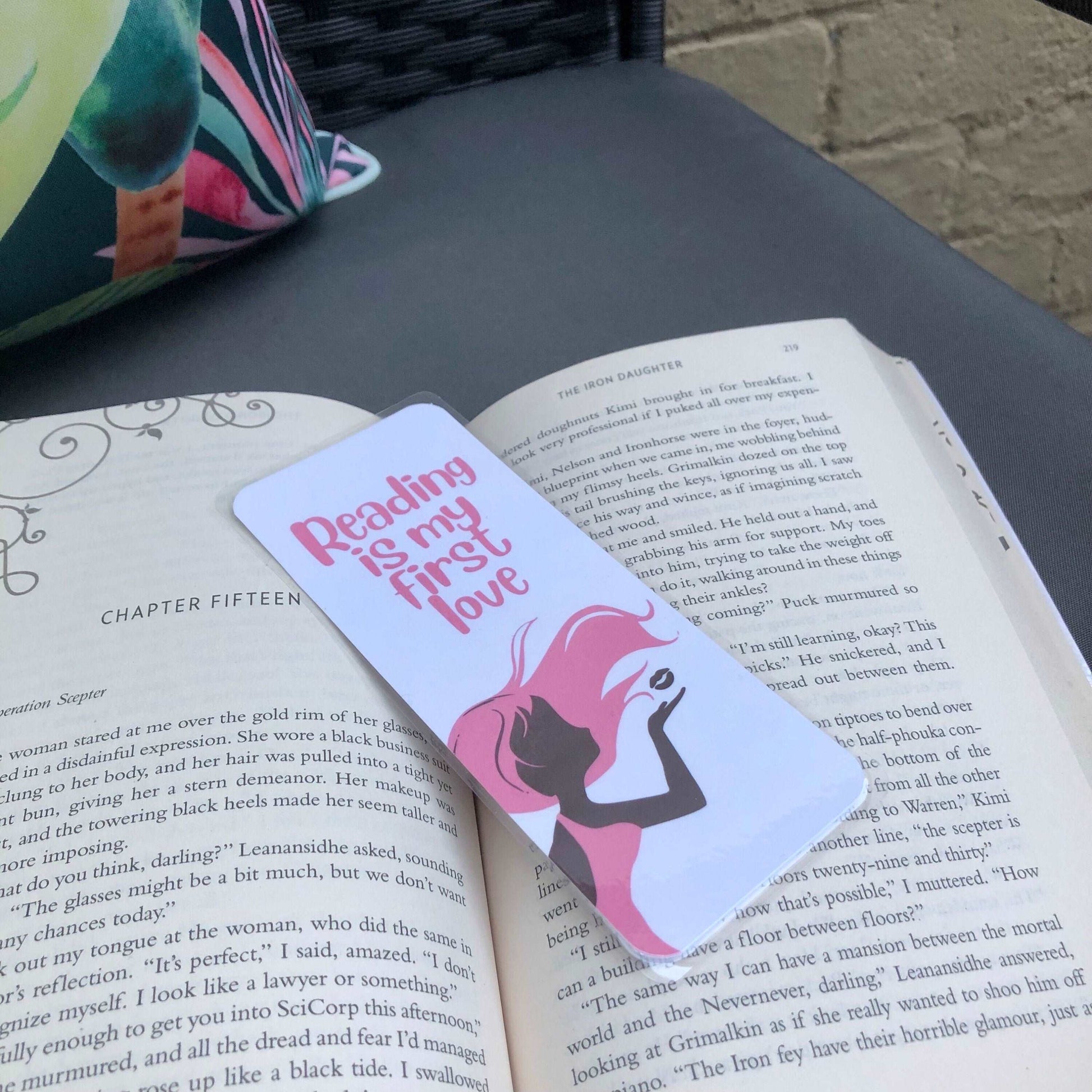 First Love Bookmark - Diamond Candlelight - girl with pink hair blowing a kiss, pink writing which says reading is my first kiss. Bookmark laying over a book sitting on a chair outside.